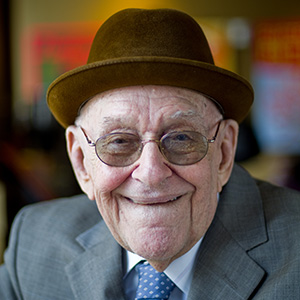 older man smiling and wearing a gray suit, brown hat and glasses